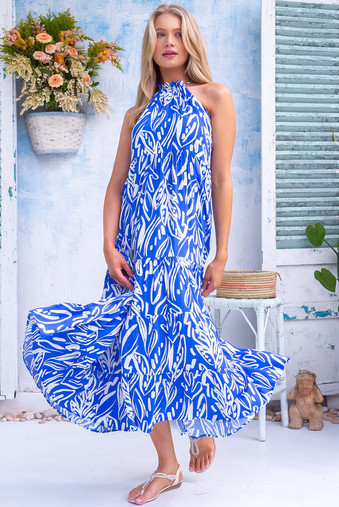 The Beach Club Ultramarine Halter Neck Maxi Dress is a beautiful blue maxi dress with a large white island print. The dress features an adjustable tie haler neckline, tiered skirting, double lined bust, side pockets, and is made from woven 100% rayon.