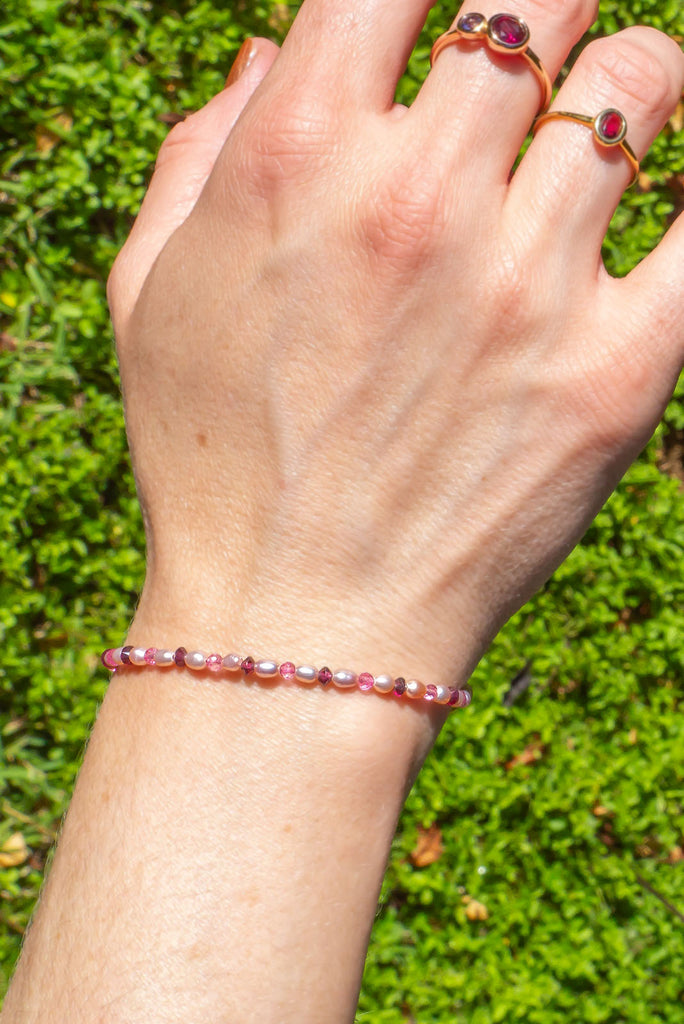 A pink pearl, pink tourmaline and rhodolite garnet delicate bracelet on a wrist with the hand showing and 2 gold rings on the fingers against a grass backdrop