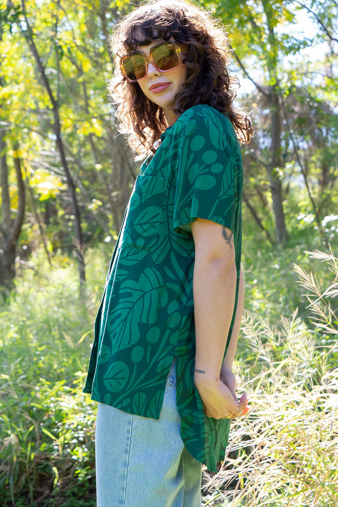 The Leonora Botany Green Short Sleeve Shirt is a tonal green relaxed fit button down shirt with botanical leaf print. The blouse features short sleeves, button down front, classic collar, one bust pocket, side slits, scooped hem at sides and slightly longer back. Made from woven 100% rayon.