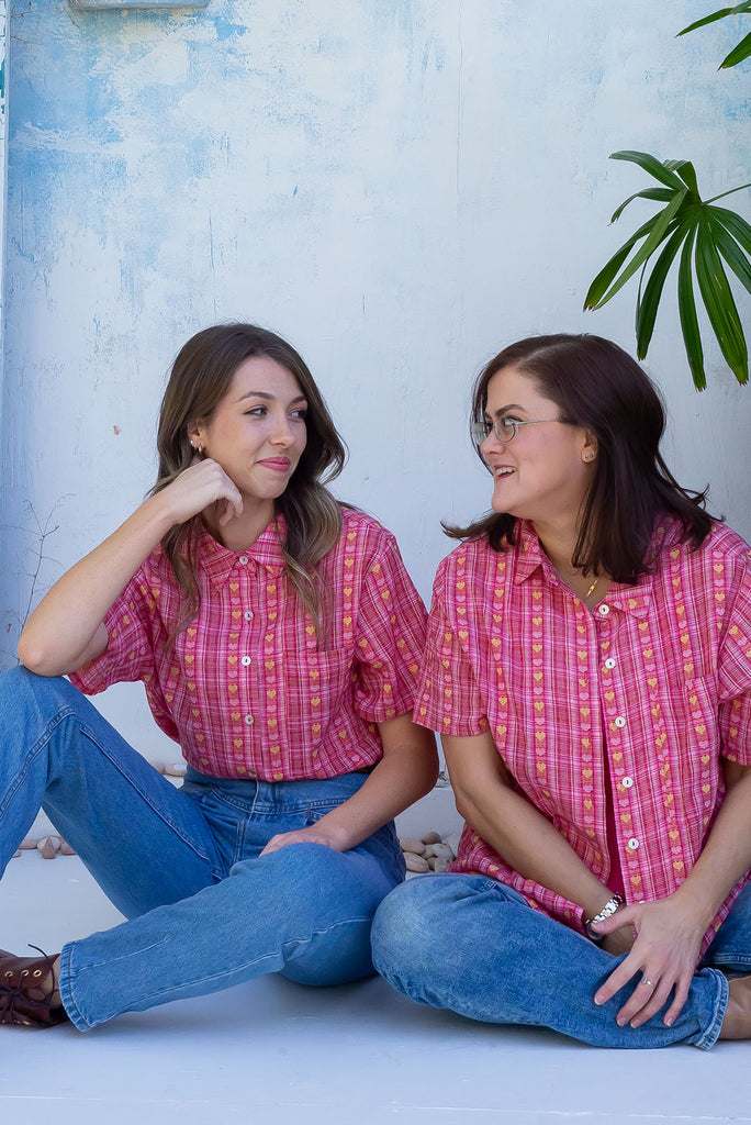 The Leonora Botany Green Short Sleeve Shirt is a plaid pink, relaxed fit, button down shirt with embroidered hearts in pink and orange. The blouse features short sleeves, button down front, classic collar, one bust pocket, side slits, scooped hem at sides and slightly longer back. Made from woven blend of cotton and polyester. 