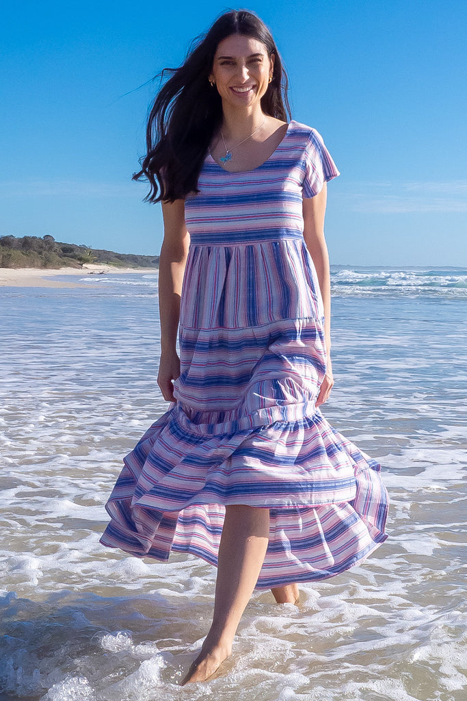 The Louisa Soft Light Stripes Maxi Dress is a beautiful tiered maxi dress with blue, white, and pink stripes. This dress features a scooped neckline, adjustable waist tabs, side pockets and a wide tiered skirt. Made from a woven blend of cotton and polyester.