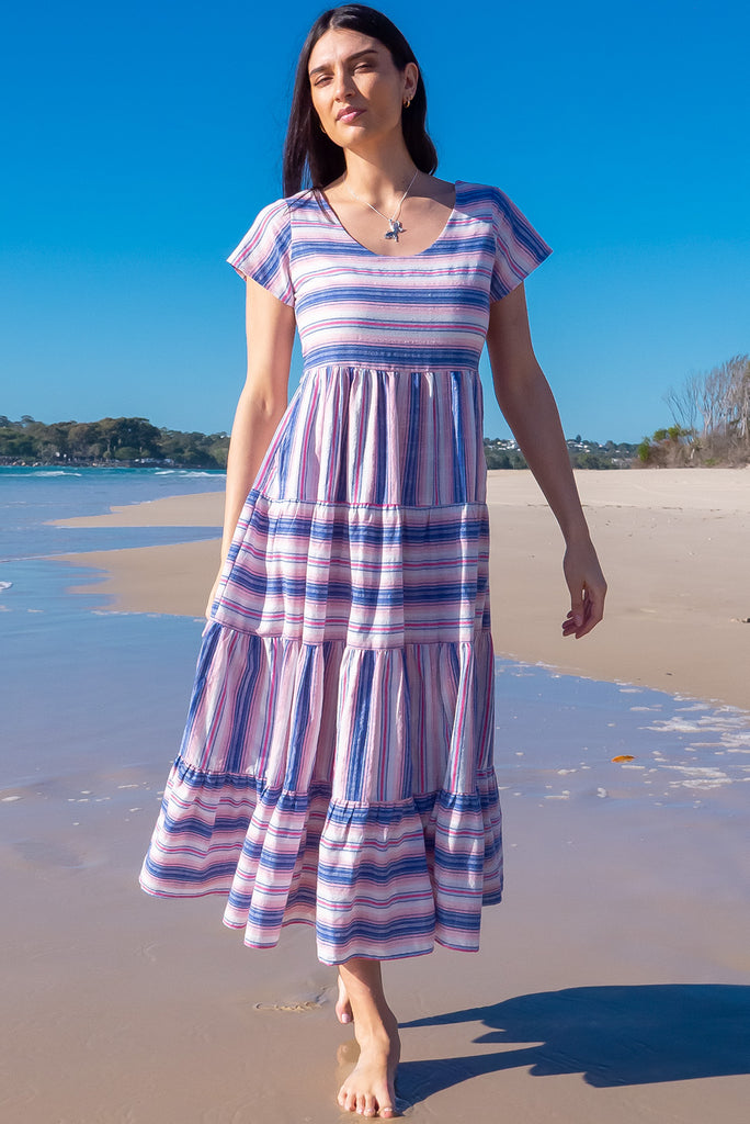 The Louisa Soft Light Stripes Maxi Dress is a beautiful tiered maxi dress with blue, white, and pink stripes. This dress features a scooped neckline, adjustable waist tabs, side pockets and a wide tiered skirt. Made from a woven blend of cotton and polyester.