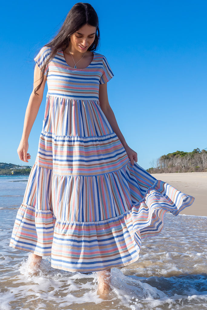 The Louisa Blue Beach Stripes Maxi Dress is a beautiful tiered maxi dress with blue, white, brown, pink and golden lurex stripes. This dress features a scooped neckline, adjustable waist tabs, side pockets and a wide tiered skirt. Made from a woven blend of cotton.