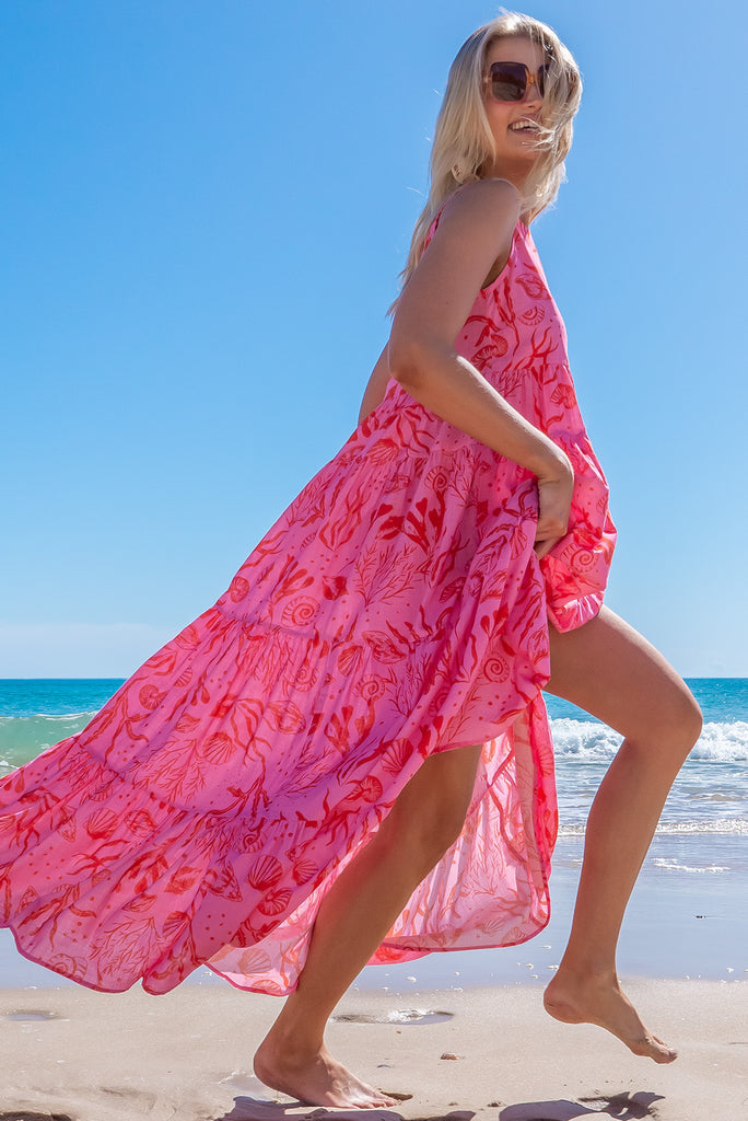 The Lulu Darling Sea Life Pink Maxi Dress features a high neck with thin straps, high cut under arms, full tiered skirting from the bust down and deep side pockets. The pink based dress has a large, red sea shell print all over. White spots may appear in the print due to the texture of the fabric. Made from 100% cotton.