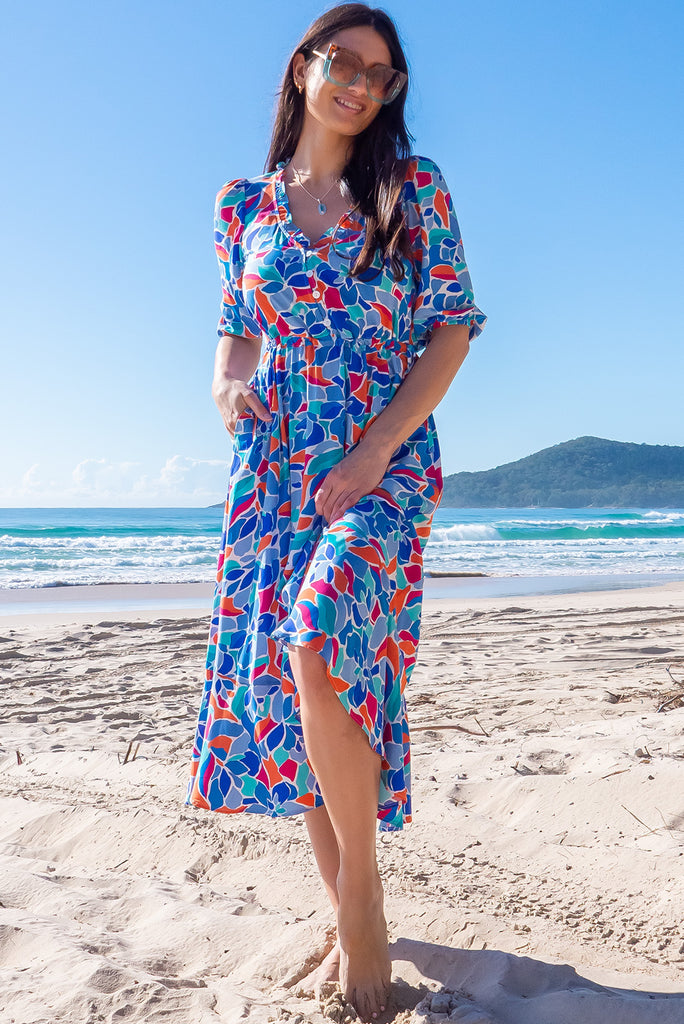 The Camellia Athens Blue Midi Dress is a beautiful blue midi dress with a multicoloured floral mosaic style print. The dress features functional buttons from front chest to waist, adjustable drawstring at either side of waist, frill v-neckline, 3/4 sleeves, and side pockets. Made from woven rayon.