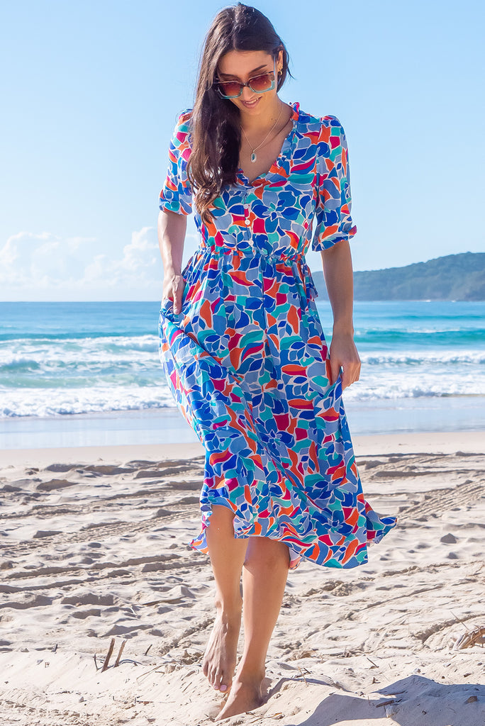 The Camellia Athens Blue Midi Dress is a beautiful blue midi dress with a multicoloured floral mosaic style print. The dress features functional buttons from front chest to waist, adjustable drawstring at either side of waist, frill v-neckline, 3/4 sleeves, and side pockets. Made from woven rayon.