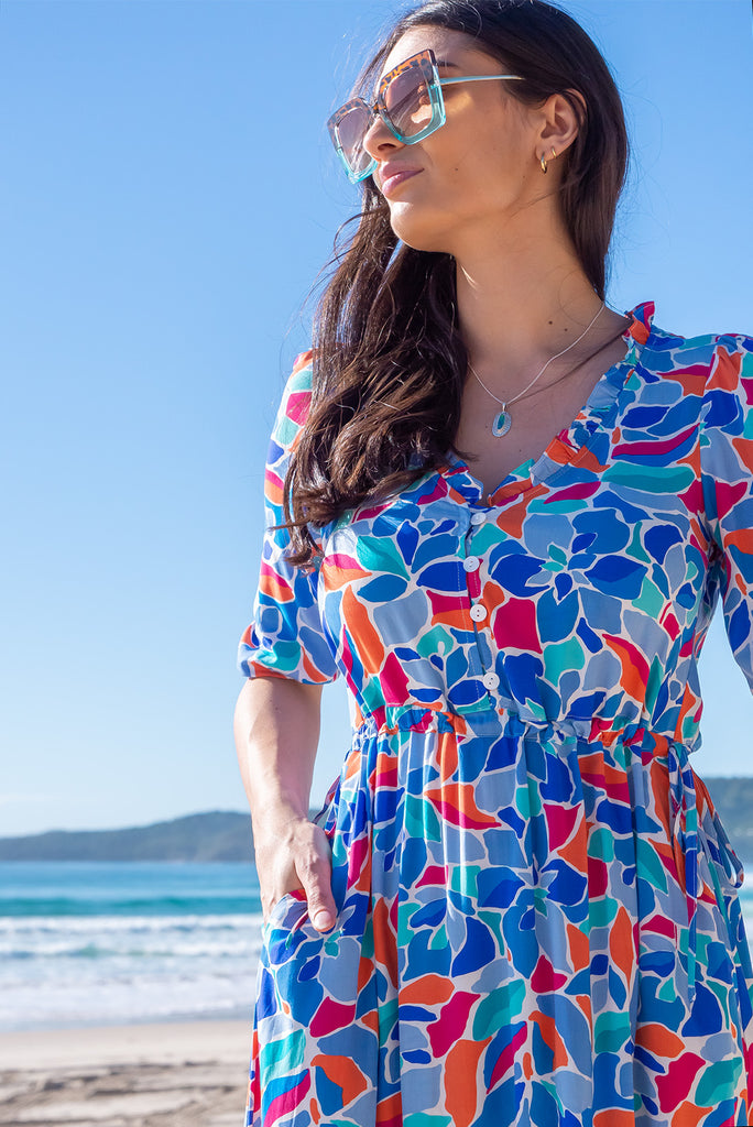 The Camellia Athens Blue Midi Dress is a beautiful blue midi dress with a multicoloured floral mosaic style print. The dress features functional buttons from front chest to waist, adjustable drawstring at either side of waist, frill v-neckline, 3/4 sleeves, and side pockets. Made from woven rayon.