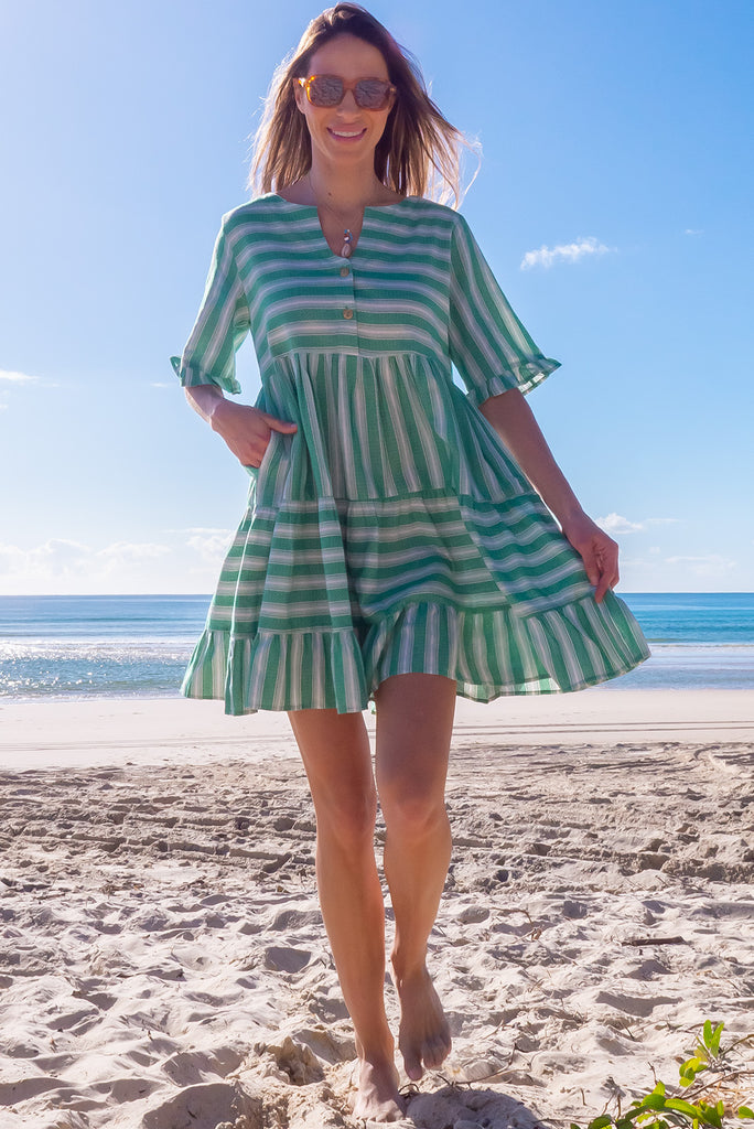 The Charlise Green Leaf Stripe Mini Dress is a green and white striped mini dress. The dress features short sleeves with frill detailing on the cuff, a split neckline with function button down chest to waistline, tiered skirting from the waist down, and side pockets. Made from woven cotton and polyester.