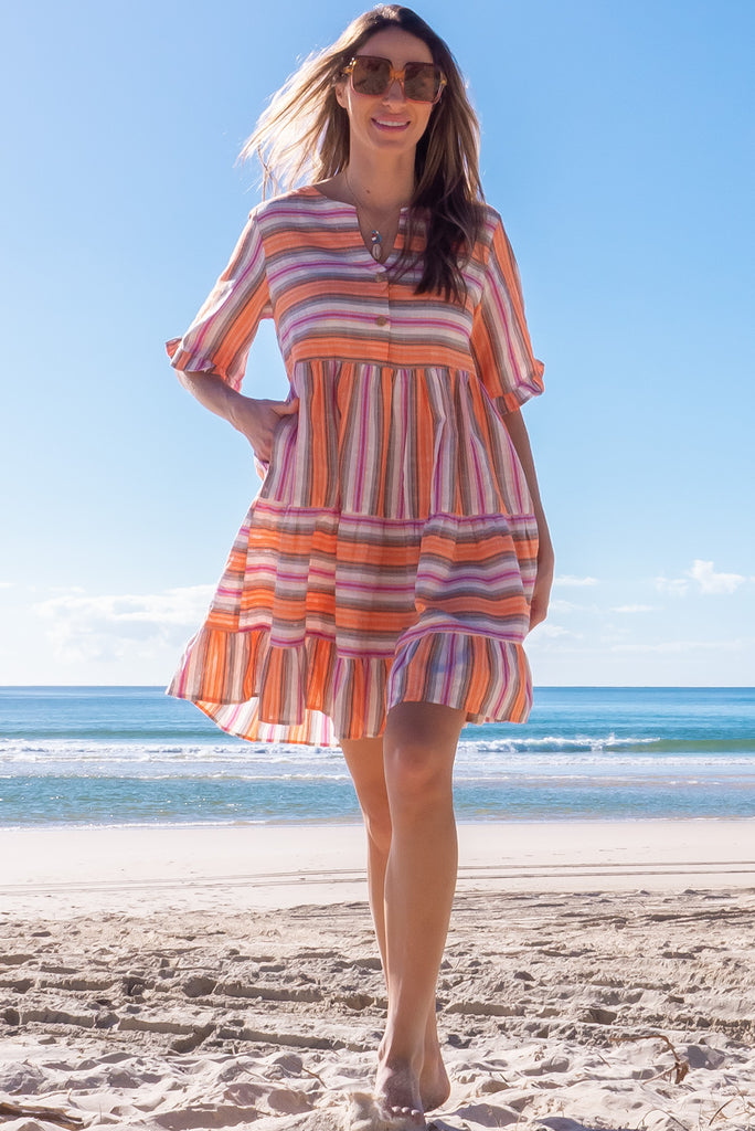 The Charlise Tuscan Stripe Mini Dress is a orange, pink, black and white striped mini dress. The dress features short sleeves with frill detailing on the cuff, a split neckline with function button down chest to waistline, tiered skirting from the waist down, and side pockets. Made from woven cotton and polyester.