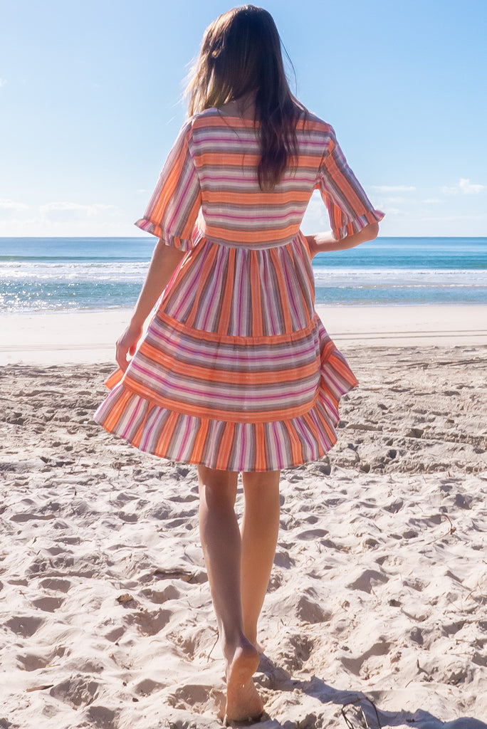 The Charlise Tuscan Stripe Mini Dress is a orange, pink, black and white striped mini dress. The dress features short sleeves with frill detailing on the cuff, a split neckline with function button down chest to waistline, tiered skirting from the waist down, and side pockets. Made from woven cotton and polyester.