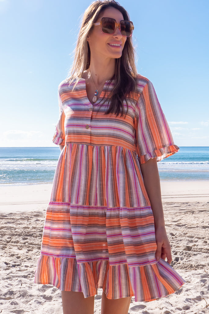 The Charlise Tuscan Stripe Mini Dress is a orange, pink, black and white striped mini dress. The dress features short sleeves with frill detailing on the cuff, a split neckline with function button down chest to waistline, tiered skirting from the waist down, and side pockets. Made from woven cotton and polyester.