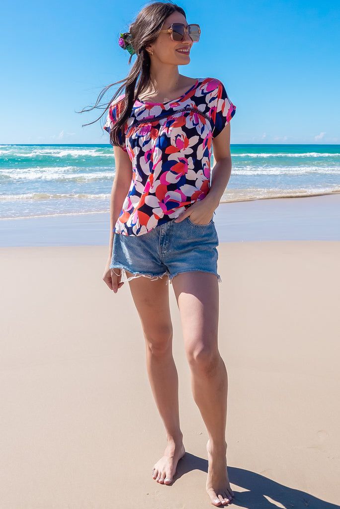 The Day Tripper Bold Summer Top is a gorgeous shirt with a bold floral print in pink, orange and blues. The top features a scooped neckline, lace insert across the chest, cap sleeves, and a small split in the side seams. Made from woven rayon.