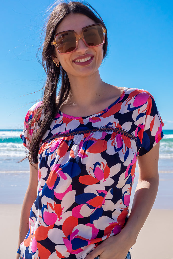 The Day Tripper Bold Summer Top is a gorgeous shirt with a bold floral print in pink, orange and blues. The top features a scooped neckline, lace insert across the chest, cap sleeves, and a small split in the side seams. Made from woven rayon.