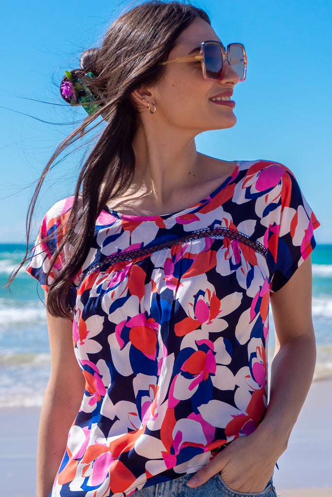 The Day Tripper Bold Summer Top is a gorgeous shirt with a bold floral print in pink, orange and blues. The top features a scooped neckline, lace insert across the chest, cap sleeves, and a small split in the side seams. Made from woven rayon.