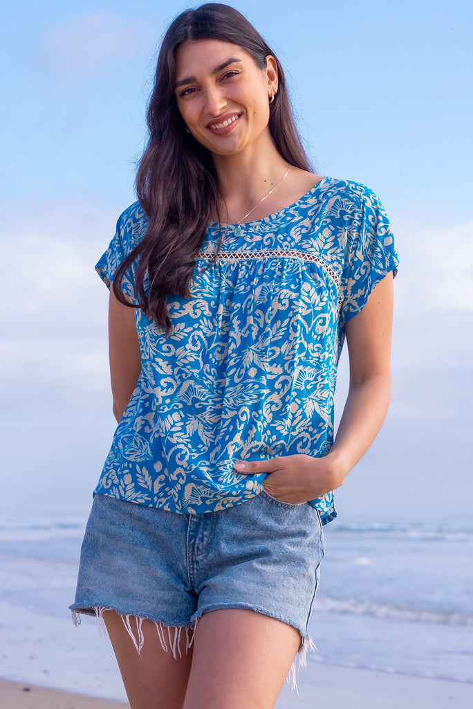 The Day Tripper Top Blue Atoll is a beautiful blue based shirt with a cream paisley print. The top features a scooped neckline, lace insert across the chest, cap sleeves, and a small split in the side seams. Made from woven rayon.