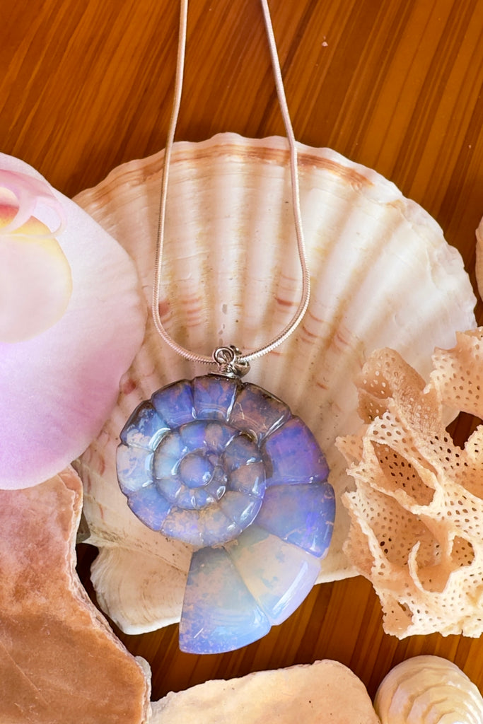 An Australian solid boulder opal pendant in the shape of a Nautilus sea shell, it has deep crystalline detail in mauve, purple and blue with slight flashes of pink. The back of the heart is polished boulder stone.