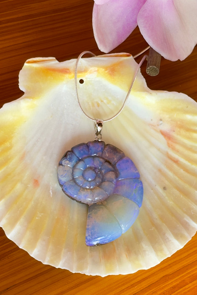 An Australian solid boulder opal pendant in the shape of a Nautilus sea shell, it has deep crystalline detail in mauve, purple and blue with slight flashes of pink. The back of the heart is polished boulder stone.