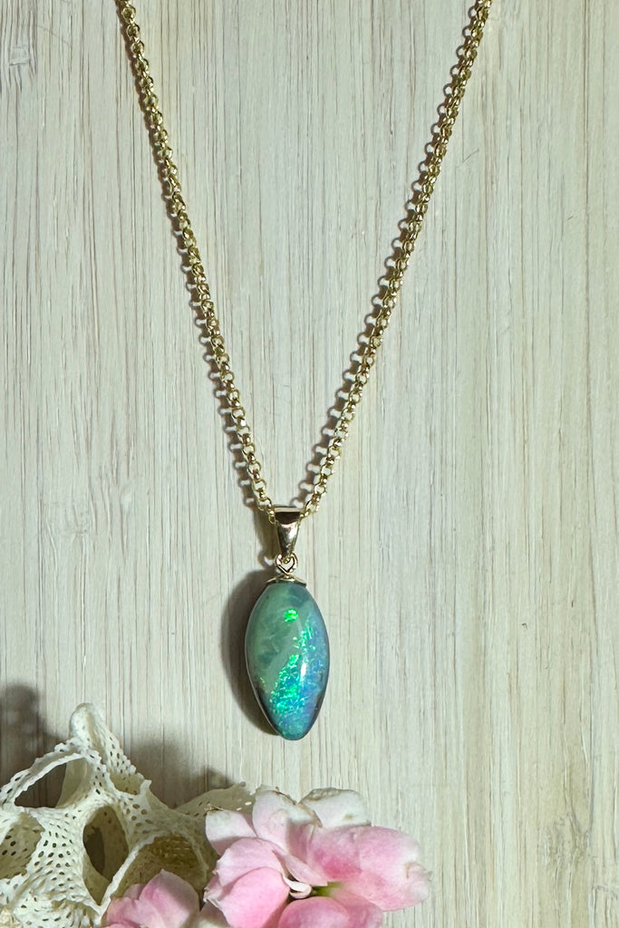 A beautiful Australian solid boulder opal pendant in the shape of a scallop shell, it has deep crystalline detail at the base, and a band of blue across the top. The natural lines of this stone appear as water flowing across the shell. 