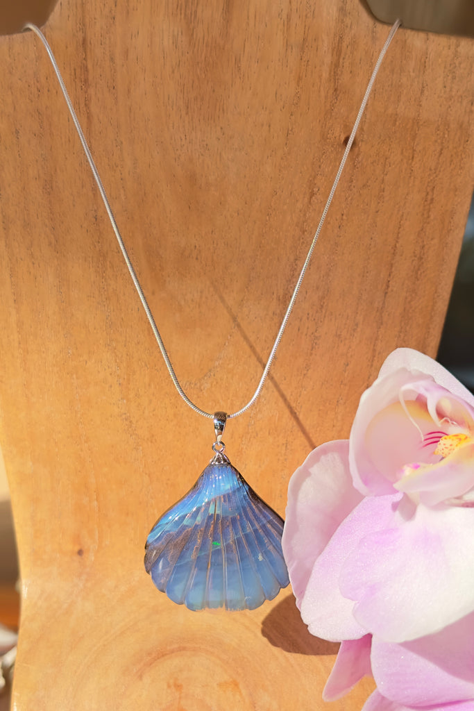 A beautiful Australian solid boulder opal pendant in the shape of a scallop shell, it has deep crystalline detail at the base, and a band of blue across the top. The natural lines of this stone appear as water flowing across the shell. 