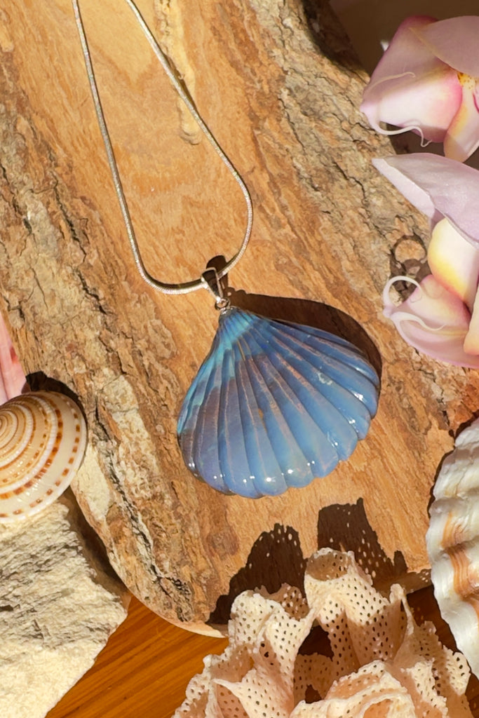 A beautiful Australian solid boulder opal pendant in the shape of a scallop shell, it has deep crystalline detail at the base, and a band of blue across the top. The natural lines of this stone appear as water flowing across the shell. 