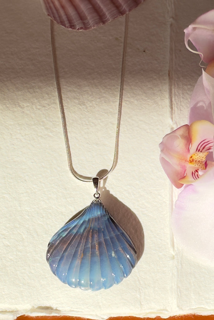 A beautiful Australian solid boulder opal pendant in the shape of a scallop shell, it has deep crystalline detail at the base, and a band of blue across the top. The natural lines of this stone appear as water flowing across the shell. 