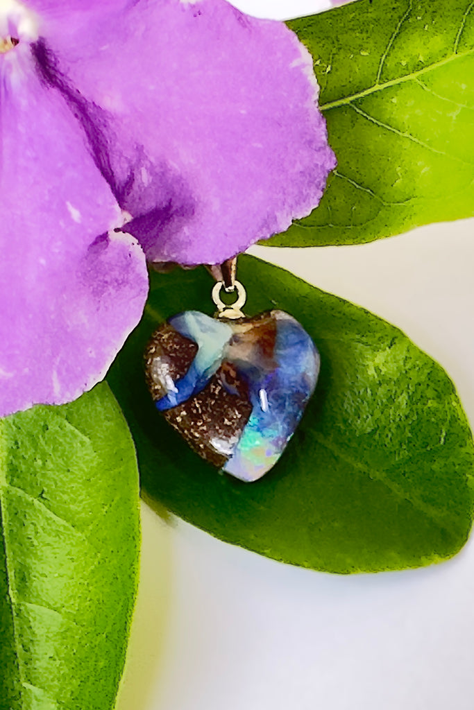 An Australian solid boulder opal pendant in a heart shape, with crystalline detail that reveals bright flashes of blue green, dark blue and misty mauve. This pretty piece has been cut into a soft heart shape, the boulder shows through in a swirling pattern across the stone. 