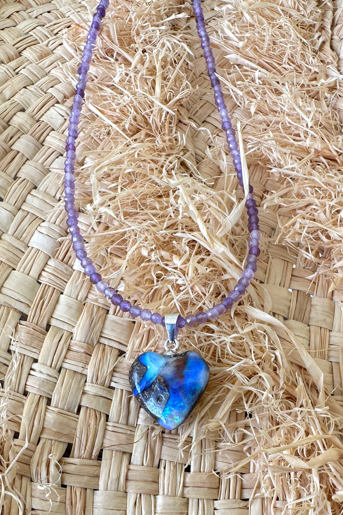 An Australian solid boulder opal pendant in a heart shape, with crystalline detail that reveals bright flashes of blue green, dark blue and misty mauve. This pretty piece has been cut into a soft heart shape, the boulder shows through in a swirling pattern across the stone. 