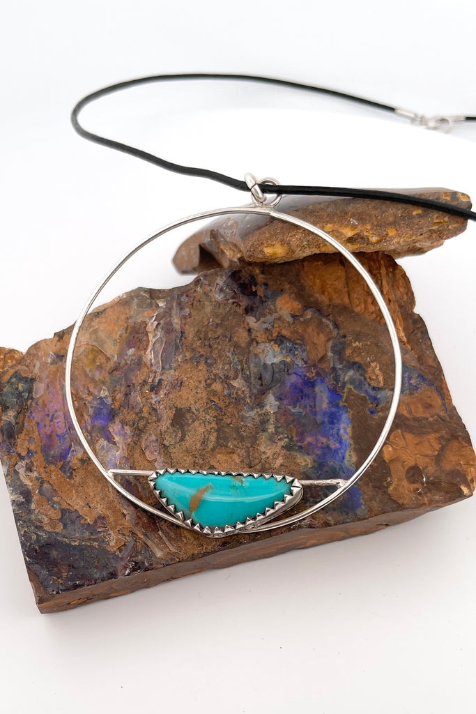 This lovely old turquoise pendant is a one off piece, hand carved into a freeform shape and showing the mark of the artist who designed it. Artisan set at the base of a hoop, it hangs from a simple black cord necklace. 