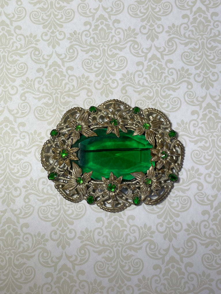 A brooch from the Edwardian era, it features a lovely emerald green centre piece and small green rhinestones that are all claw set with none missing. 