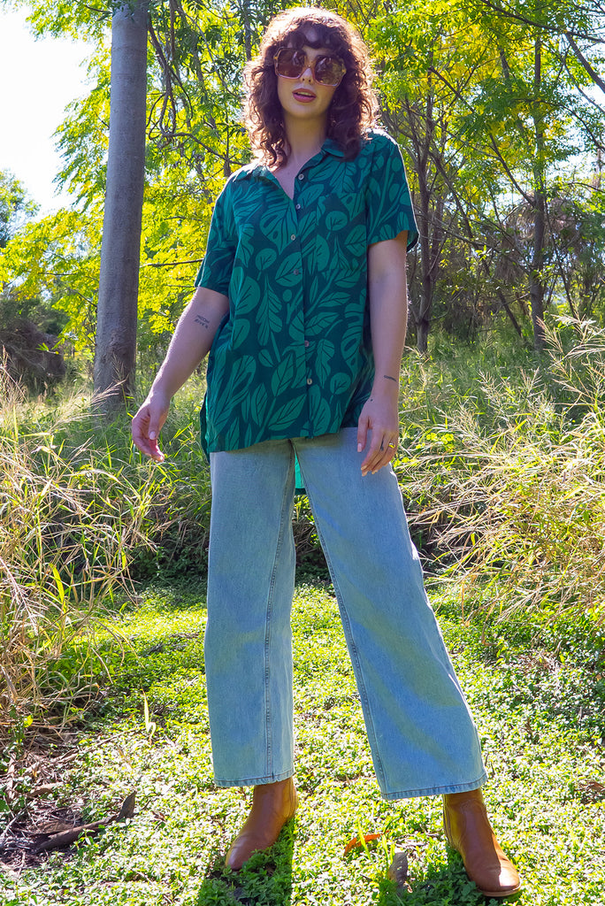 The Leonora Botany Green Short Sleeve Shirt is a tonal green relaxed fit button down shirt with botanical leaf print. The blouse features short sleeves, button down front, classic collar, one bust pocket, side slits, scooped hem at sides and slightly longer back. Made from woven 100% rayon.