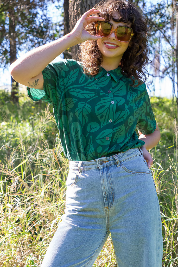 The Leonora Botany Green Short Sleeve Shirt is a tonal green relaxed fit button down shirt with botanical leaf print. The blouse features short sleeves, button down front, classic collar, one bust pocket, side slits, scooped hem at sides and slightly longer back. Made from woven 100% rayon.