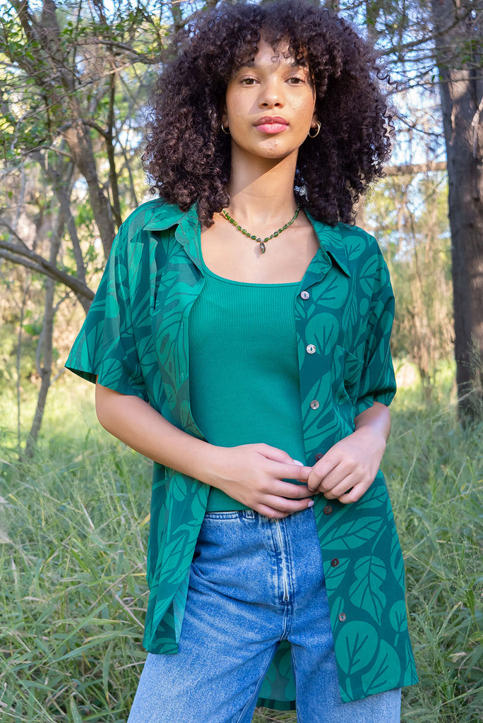 The Leonora Botany Green Short Sleeve Shirt is a tonal green relaxed fit button down shirt with botanical leaf print. The blouse features short sleeves, button down front, classic collar, one bust pocket, side slits, scooped hem at sides and slightly longer back. Made from woven 100% rayon.