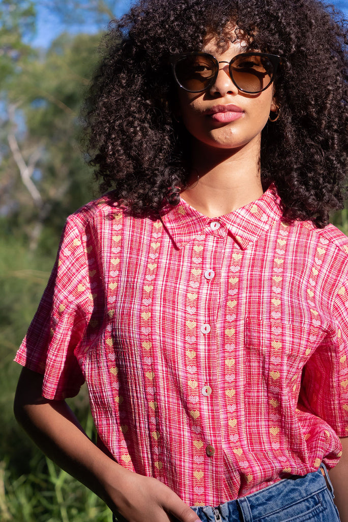 The Leonora Botany Green Short Sleeve Shirt is a plaid pink, relaxed fit, button down shirt with embroidered hearts in pink and orange. The blouse features short sleeves, button down front, classic collar, one bust pocket, side slits, scooped hem at sides and slightly longer back. Made from woven blend of cotton and polyester. 