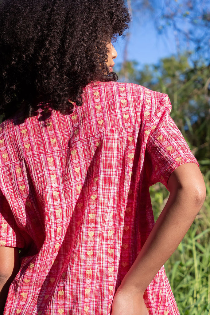 The Leonora Botany Green Short Sleeve Shirt is a plaid pink, relaxed fit, button down shirt with embroidered hearts in pink and orange. The blouse features short sleeves, button down front, classic collar, one bust pocket, side slits, scooped hem at sides and slightly longer back. Made from woven blend of cotton and polyester. 