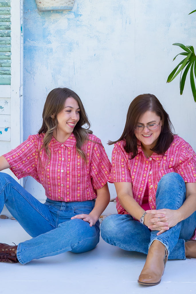 The Leonora Botany Green Short Sleeve Shirt is a plaid pink, relaxed fit, button down shirt with embroidered hearts in pink and orange. The blouse features short sleeves, button down front, classic collar, one bust pocket, side slits, scooped hem at sides and slightly longer back. Made from woven blend of cotton and polyester. 