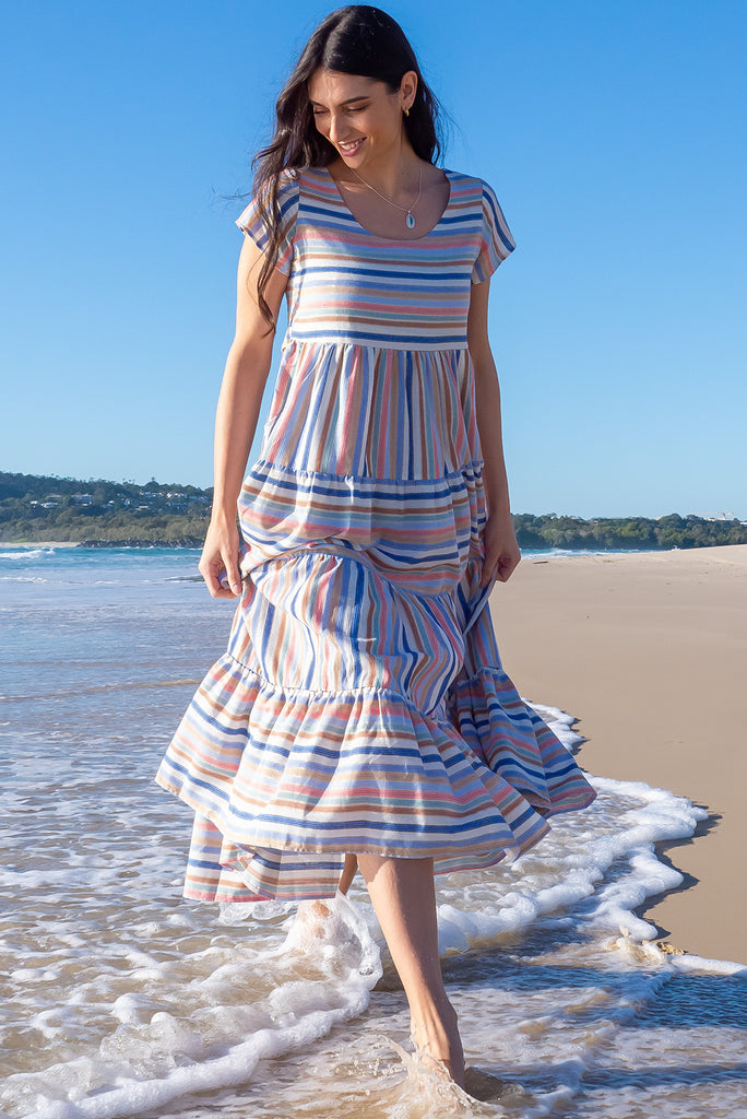 The Louisa Blue Beach Stripes Maxi Dress is a beautiful tiered maxi dress with blue, white, brown, pink and golden lurex stripes. This dress features a scooped neckline, adjustable waist tabs, side pockets and a wide tiered skirt. Made from a woven blend of cotton.