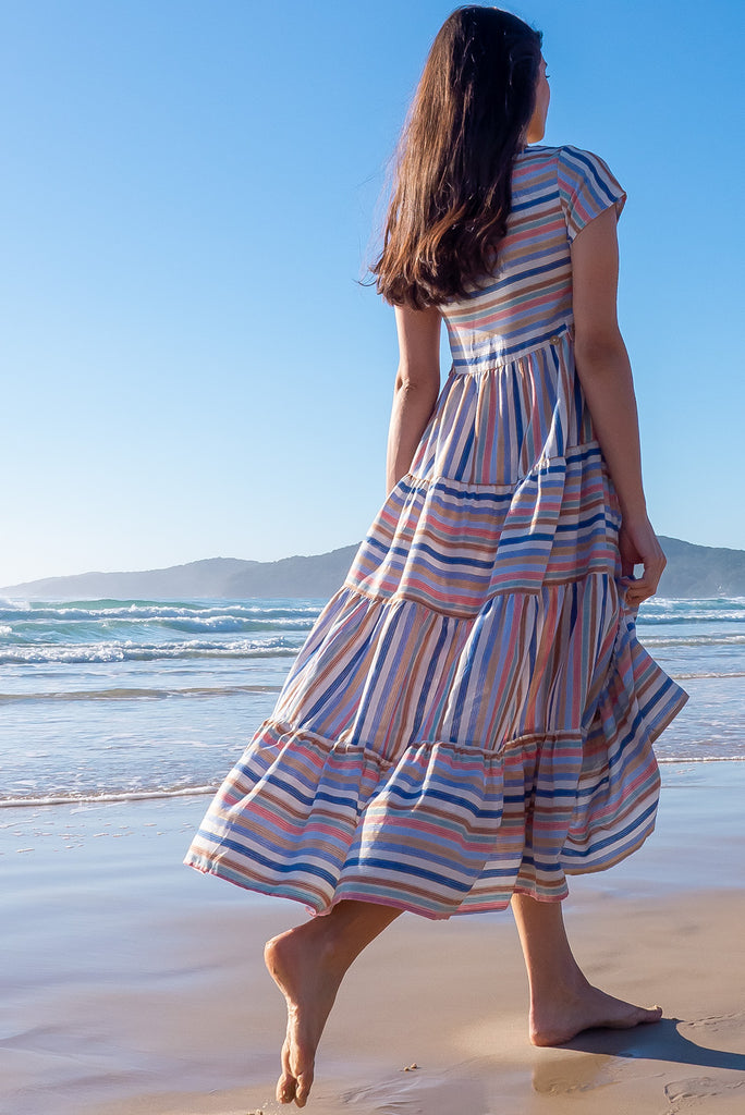 The Louisa Blue Beach Stripes Maxi Dress is a beautiful tiered maxi dress with blue, white, brown, pink and golden lurex stripes. This dress features a scooped neckline, adjustable waist tabs, side pockets and a wide tiered skirt. Made from a woven blend of cotton.