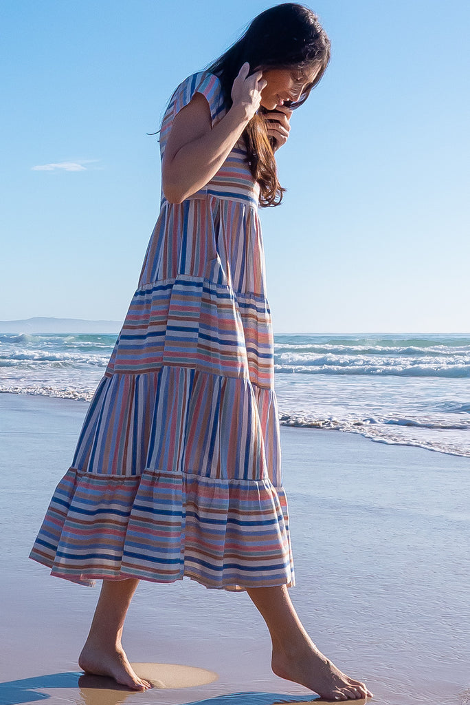 The Louisa Blue Beach Stripes Maxi Dress is a beautiful tiered maxi dress with blue, white, brown, pink and golden lurex stripes. This dress features a scooped neckline, adjustable waist tabs, side pockets and a wide tiered skirt. Made from a woven blend of cotton.