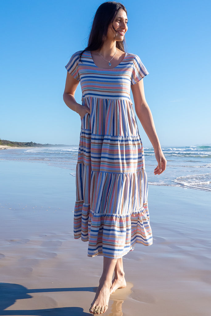 The Louisa Blue Beach Stripes Maxi Dress is a beautiful tiered maxi dress with blue, white, brown, pink and golden lurex stripes. This dress features a scooped neckline, adjustable waist tabs, side pockets and a wide tiered skirt. Made from a woven blend of cotton.