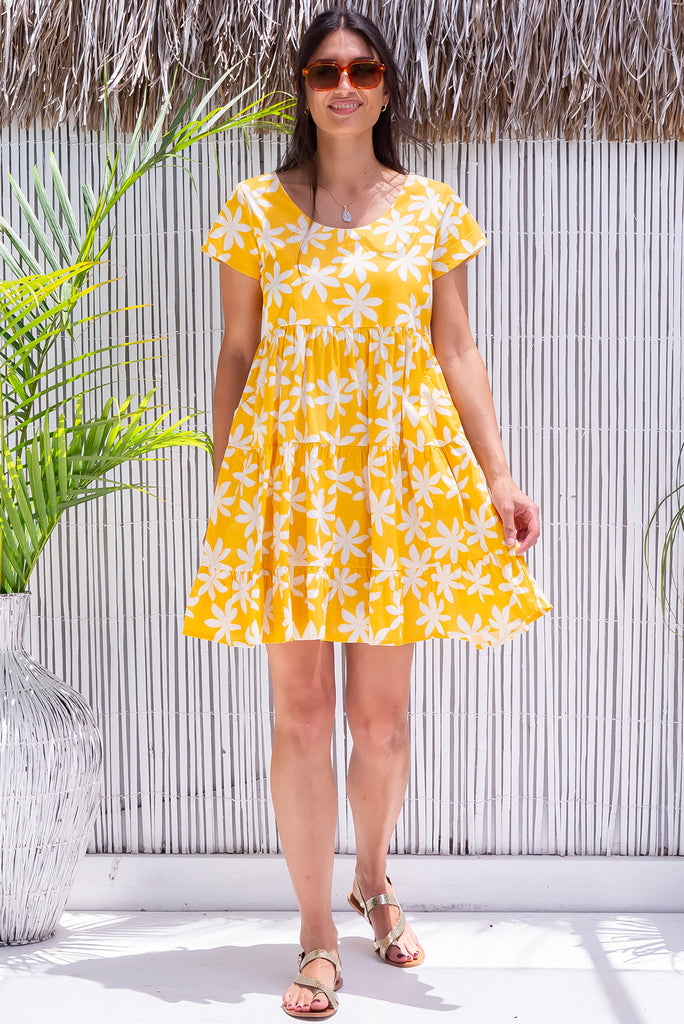 The Marini Crazy Dazy Mini Dress is a vibrant yellow mini dress with a large white floral daisy print. The dress features waist tabs at the back, short sleeves and tiered skirting. Made from 100% cotton. 