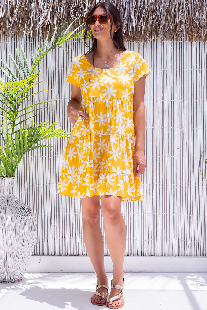 The Marini Crazy Dazy Mini Dress is a vibrant yellow mini dress with a large white floral daisy print. The dress features waist tabs at the back, short sleeves and tiered skirting. Made from 100% cotton. 