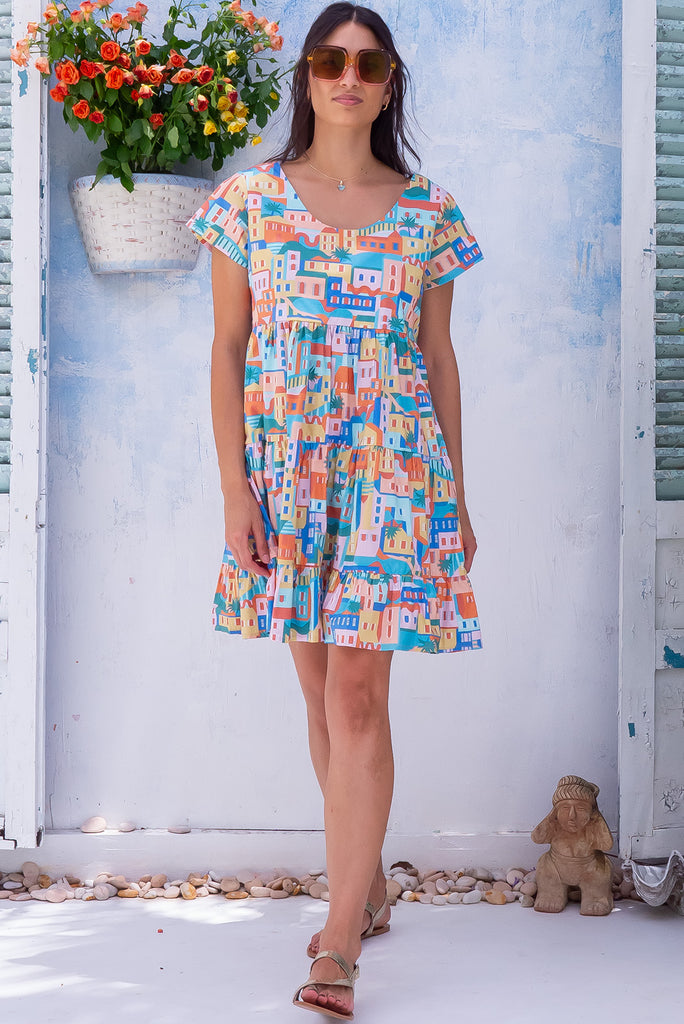 The Marini Sandcastle Mini Dress is a gorgeous mini dress with a graphic house silhouette print in pastel shades. The dress features waist tabs at the back, short sleeves and tiered skirting. Made from 100% cotton poplin. 