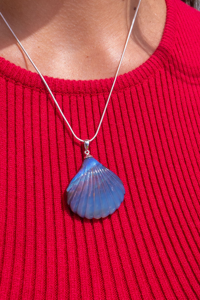 A beautiful Australian solid boulder opal pendant in the shape of a scallop shell, it has deep crystalline detail at the base, and a band of blue across the top. The natural lines of this stone appear as water flowing across the shell.