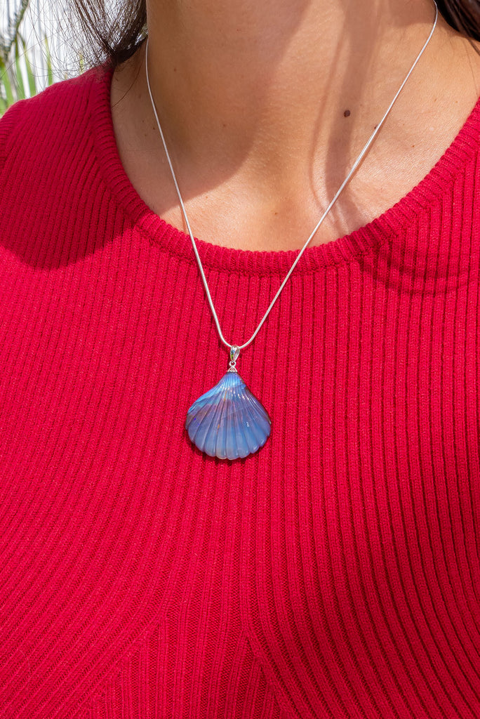A beautiful Australian solid boulder opal pendant in the shape of a scallop shell, it has deep crystalline detail at the base, and a band of blue across the top. The natural lines of this stone appear as water flowing across the shell.