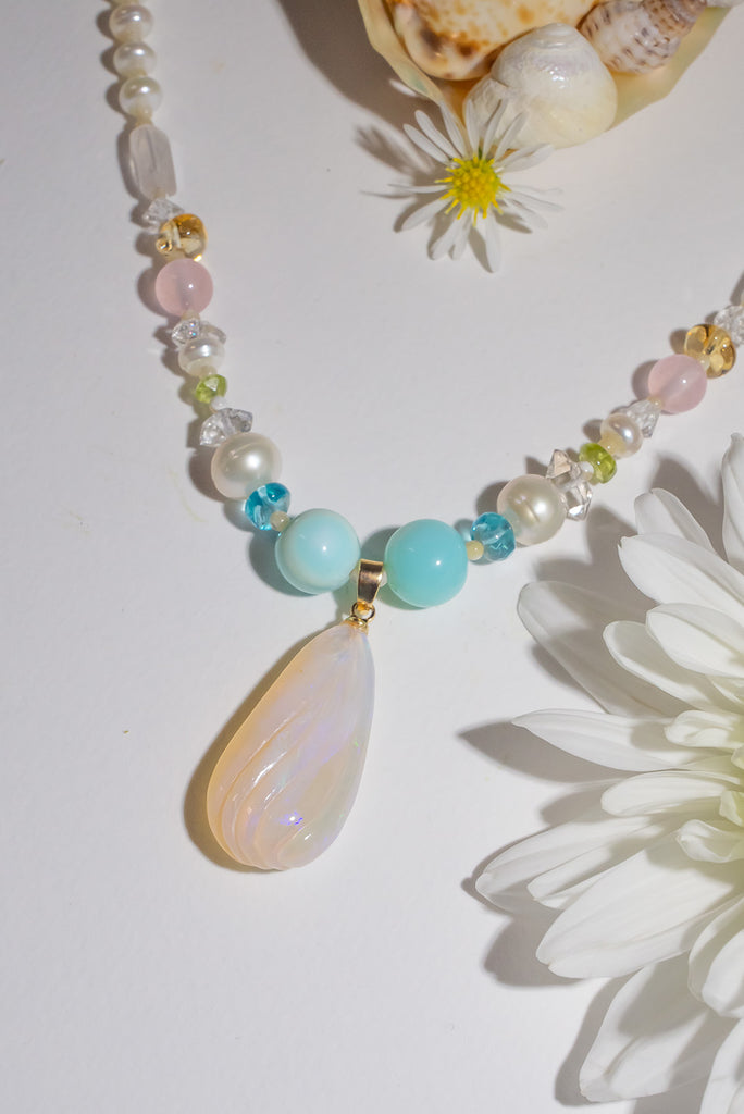 A beautiful creamy pink hand carved crystal opal with subtle flashes of green and blue, on a necklace of small lustrous pearls with gemstone highlights.