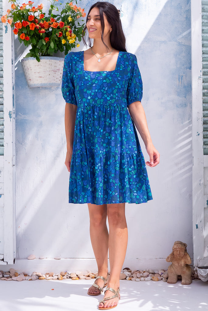 The Rosie Sea Shells Blue Mini Dress is a beautiful deep blue mini dress with a small cool-toned seashell print all over. The dress features a square neckline, puffy sleeves,  and adjustable waist tabs at the back. Made from a woven blend of cotton and rayon.