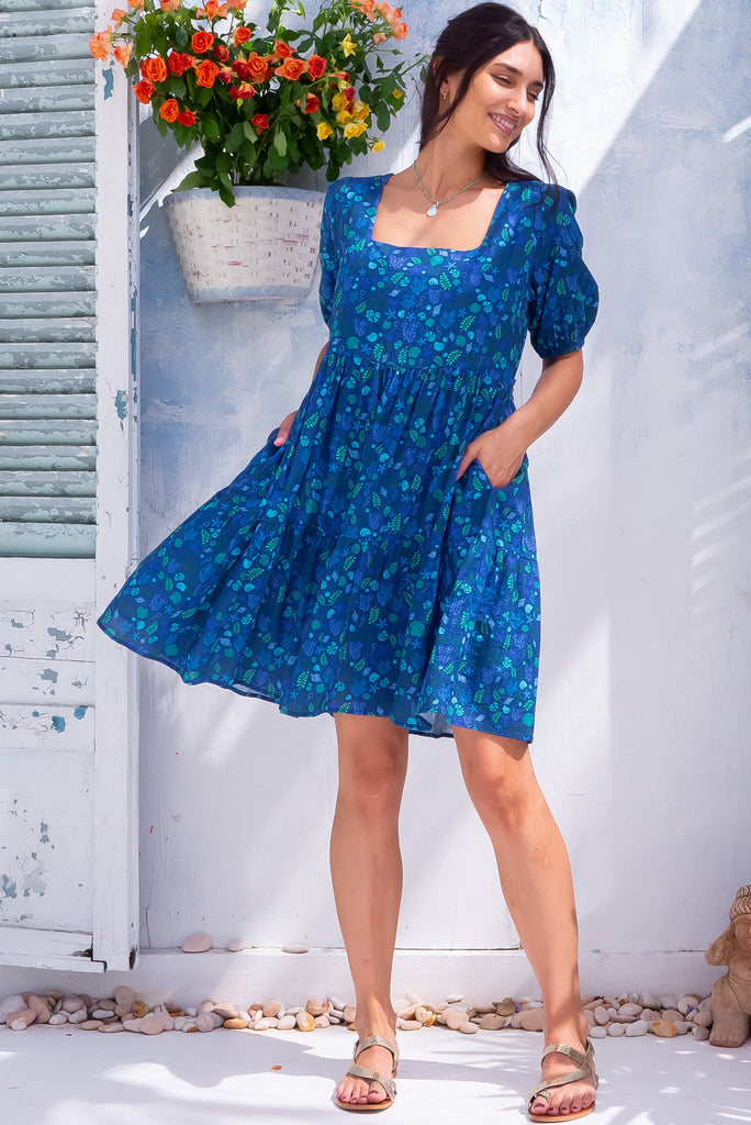 The Rosie Sea Shells Blue Mini Dress is a beautiful deep blue mini dress with a small cool-toned seashell print all over. The dress features a square neckline, puffy sleeves,  and adjustable waist tabs at the back. Made from a woven blend of cotton and rayon.