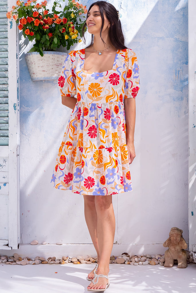 The Rosie Mediterranean Flow Mini Dress is a beautiful off-white based mini dress with a warm toned midetemrranean style print. The dress features a square neckline, puffy sleeves,  and adjustable waist tabs at the back. Made from a woven blend of cotton and rayon.