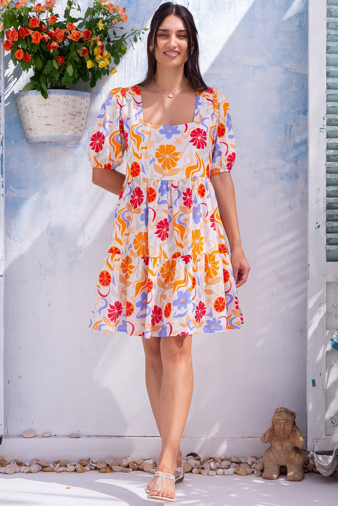 The Rosie Mediterranean Flow Mini Dress is a beautiful off-white based mini dress with a warm toned midetemrranean style print. The dress features a square neckline, puffy sleeves,  and adjustable waist tabs at the back. Made from a woven blend of cotton and rayon.