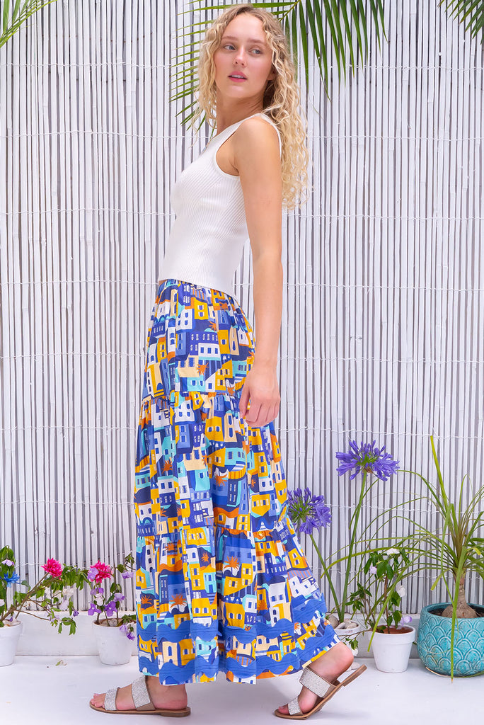 The Sorrento Sandcastles Maxi Skirt is a gorgeous skirt with a graphic house silhouette print in shades of yellow and blue. The skirt features a slip on design, elasticated waistband, side pockets and is tiered with a blue oversized ric-rac braid feature. Made from 100% cotton poplin.
